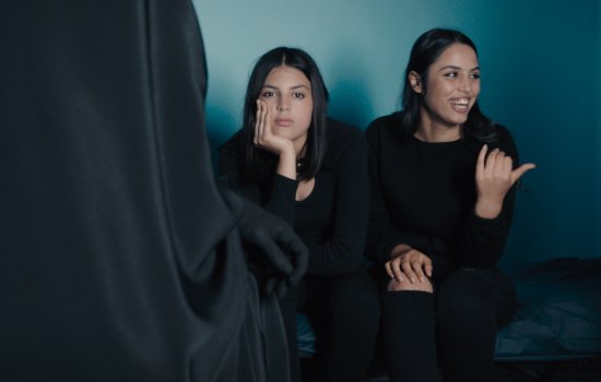 Two women are sat. One stares straight into the camera as another smiles and gestures out of frame.