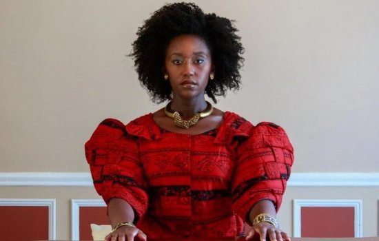 A woman in a vibrant red dress is sat staring directly into the camera.