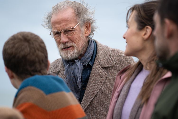 An older man with glasses, a tweed jacket and blue scarf looks at a boy. Next to the boy are a woman and a man.