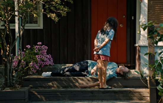 Promotional image for Blue Heron; A young girl wearing a blue tshirt and red shorts stands looking at a woman who is lying on a porch.