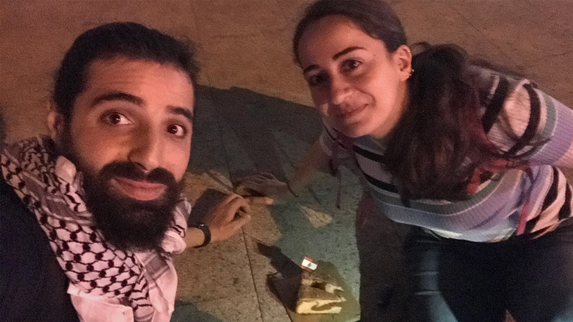Promotional image from Birds of War; a man and a woman take a selfie, they are sitting on the ground, between them is a peice of cake.