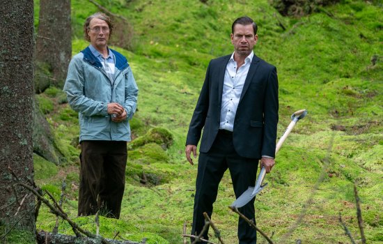 Promotional image for The Last Viking; A man in a blue coat and a man in a suit stand in the woods. The man in the suit is holding a shovel.