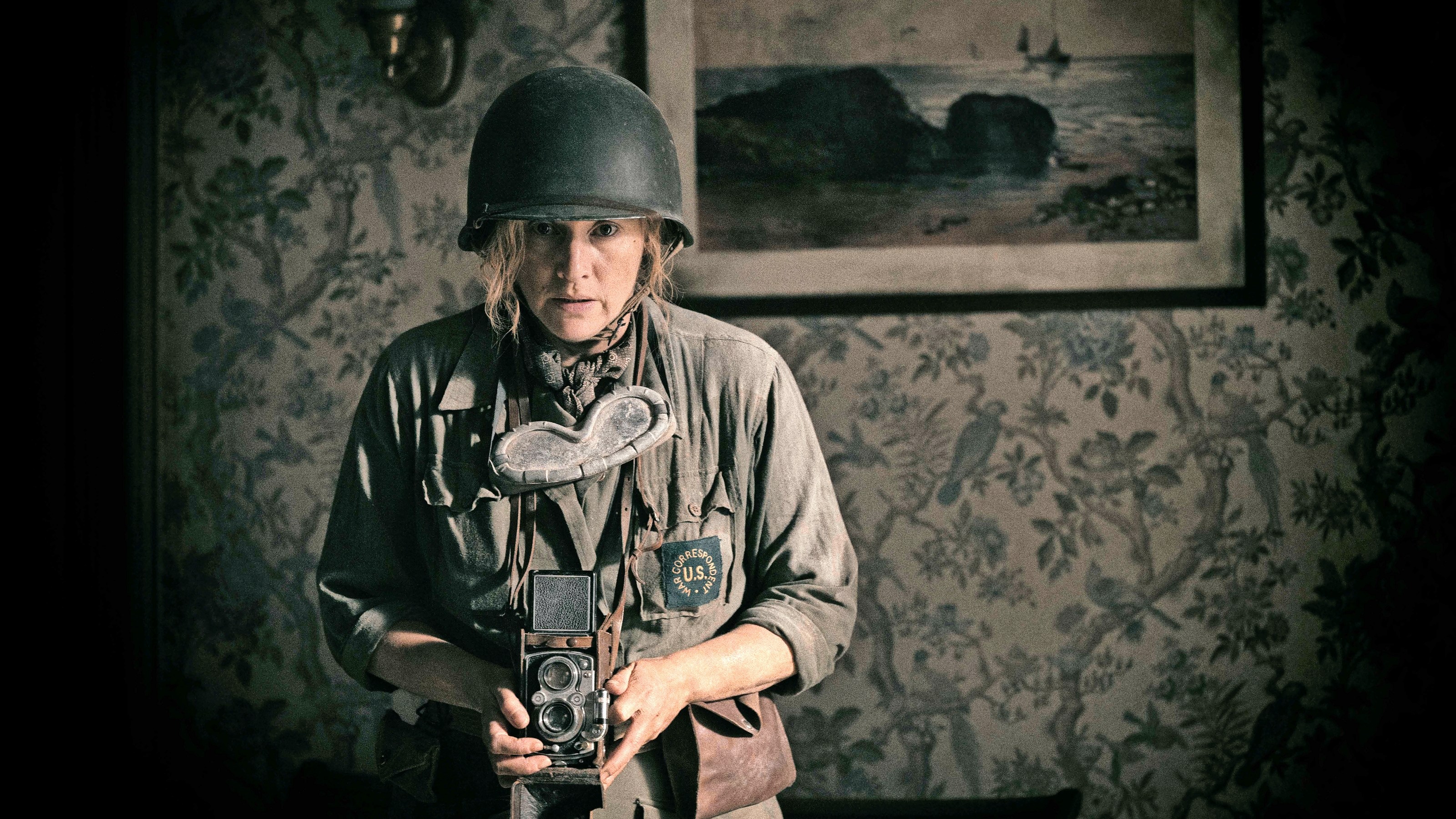 Kate Winslet points a camera at a subject offscreen. She is dressed in army attire.