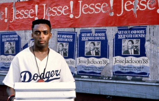 Spike Lee stands in front of a colourful backdrop holding a pizza box.