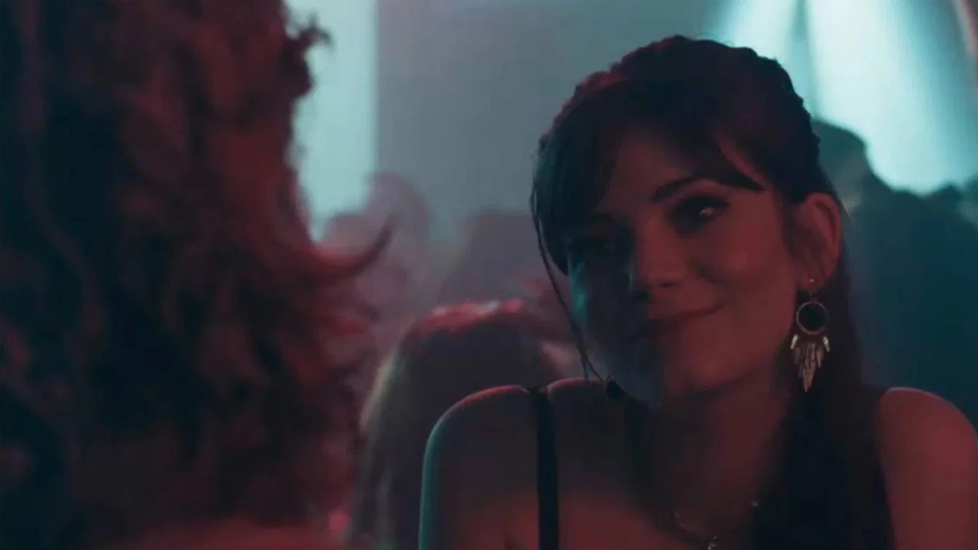 A woman with long dark hair and dangly earrings smiles at another person who is off camera