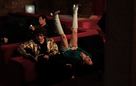 A woman sits upside down on a chair as a man reads a book next to her.