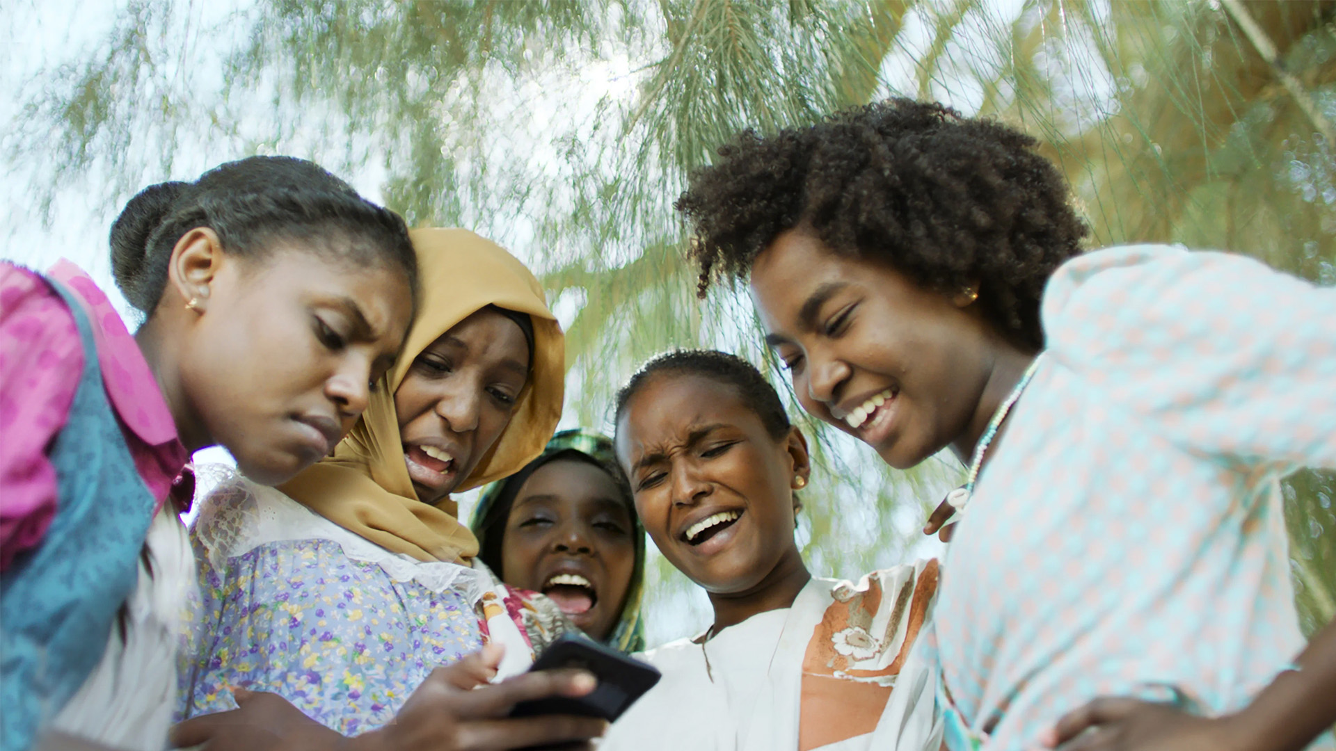 A group of young girls huddle around a phone, laughing