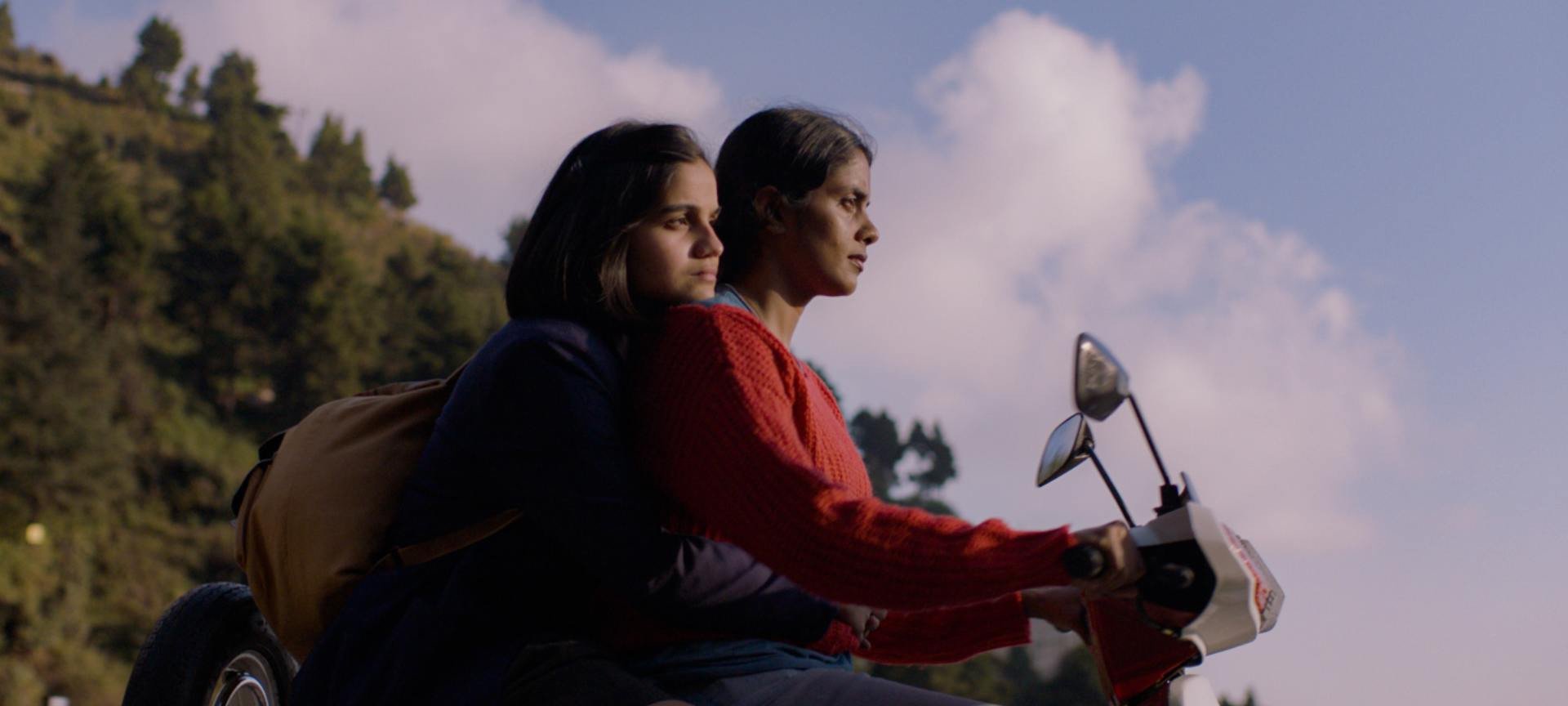 Two women ride a motorcycle together, with a cloudy backdrop behind them.