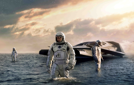 A group of people in spacesuits stand in the middle of water, their spaceship sits in the background.