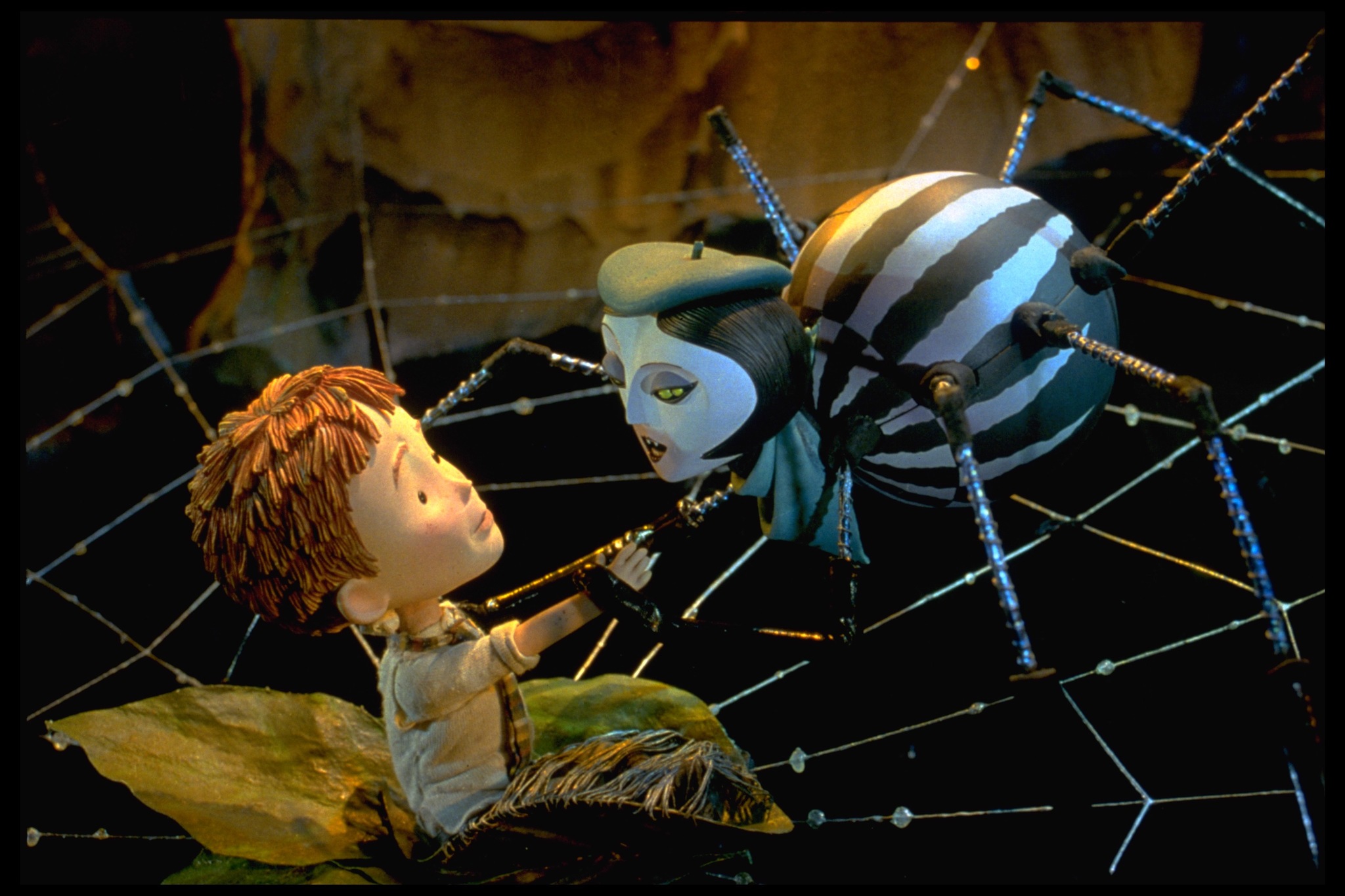 A stop-motion image of a young boy sat on a large spider web as a spider wearing a beret grabs his arm.