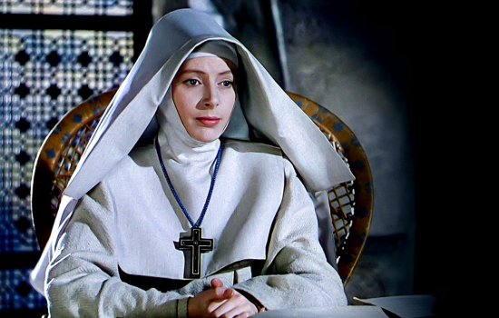 A nun sits in a wicker chair, wearing a large cross.