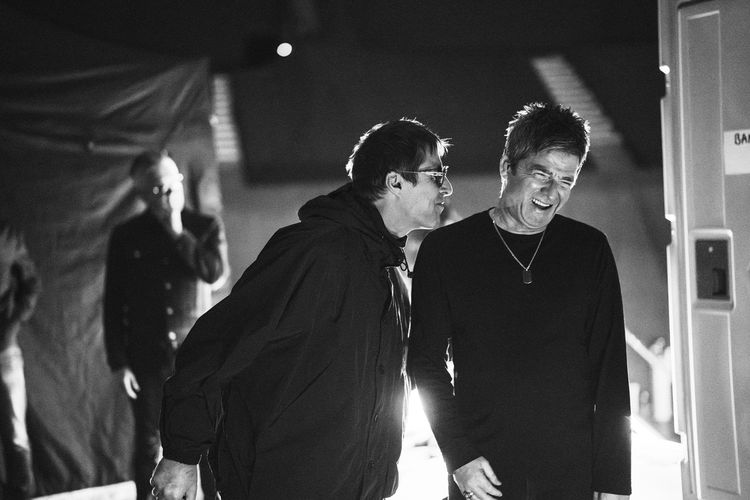 A black and white image of Liam Gallagher leaning in to speak to Noel Gallagher, who laughs.