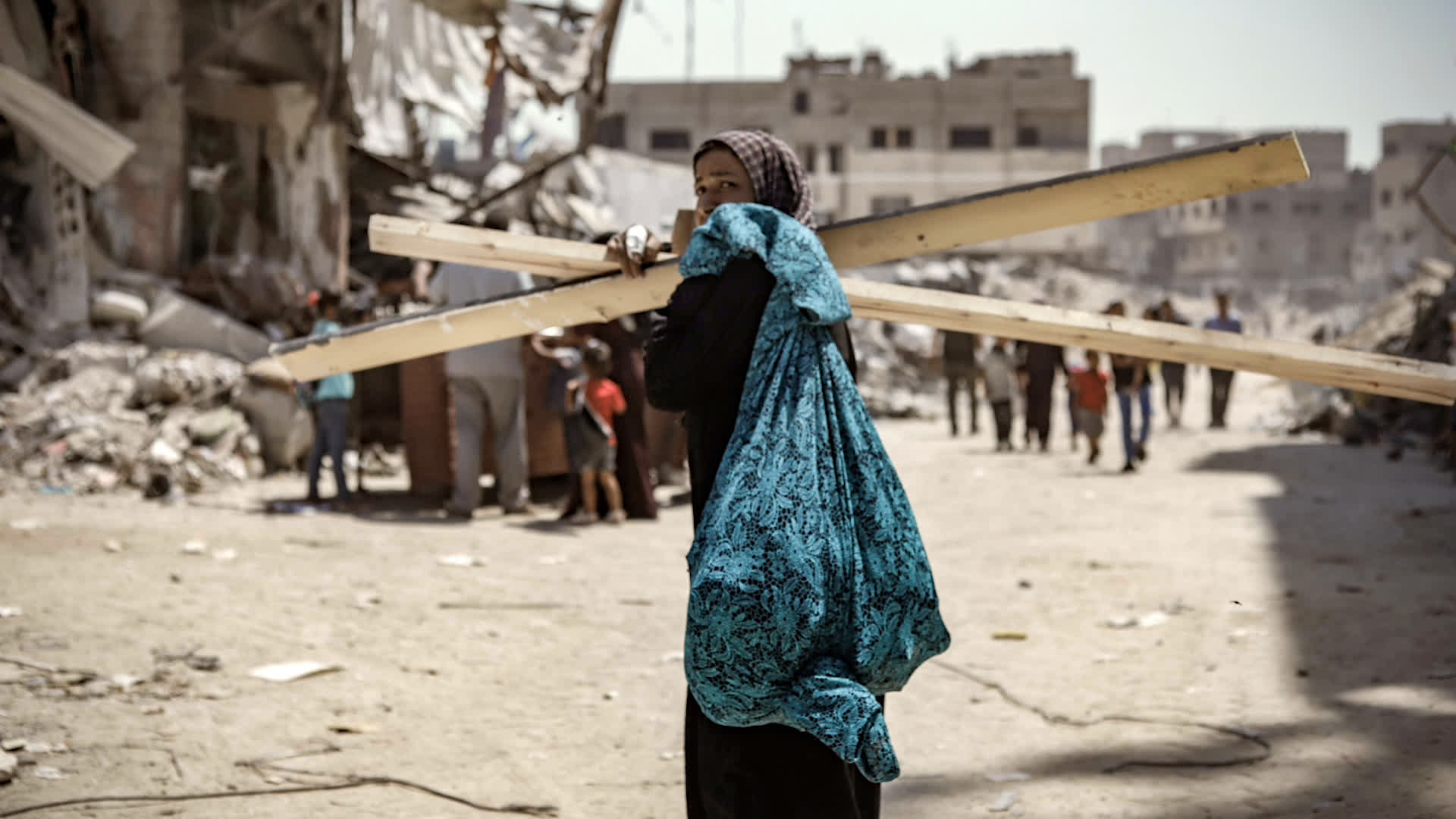 A woman works through a street covered in rubble, she carries some planks of wood/