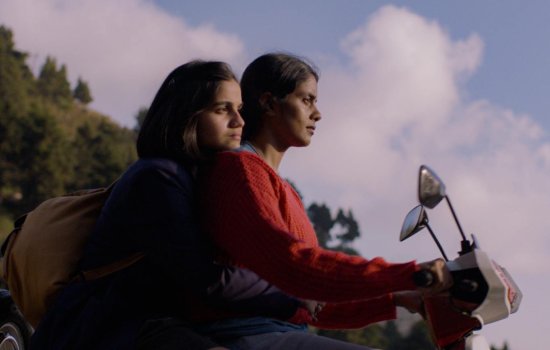 Two women ride a motorcycle together, with a cloudy backdrop behind them.