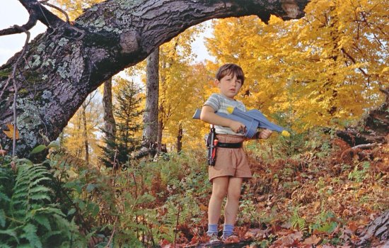 A young boy stands in a colourful, leafy setting holding a toy gun.