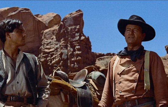 John Wayne wears a cowboy hat and stands in a cavernous location.