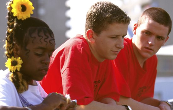 Two young men and a a young woman stand in a row. The men are both dressed in bright red t-shirts while the woman wears sunflowers in her hair.
