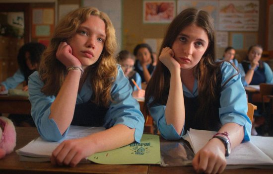 Two girls sit side by side in a classroom, their heads resting on their hands.