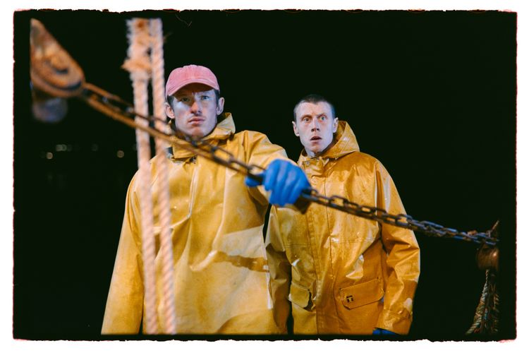 Callum Turner and George Mackay are dressed in yellow fisherman's coats, standing against the black night.