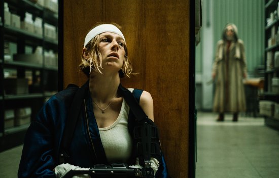 Hunter Schafer hides behind some shelves with a bandaged head, as a shadowy figure looks for her in the distance.