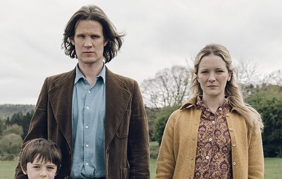 Matt Smith, Morfydd Clark and a young boy stand side by side in a grey field, wearing 70s clothing and with blank expressions on their faces.