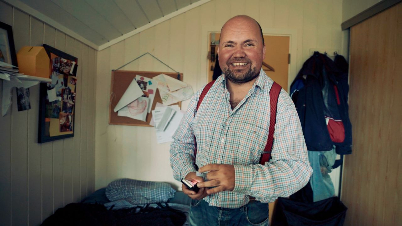 A man smiles and looks at the camera while stood in a bedroom. He wears a checked shirt with red suspenders.