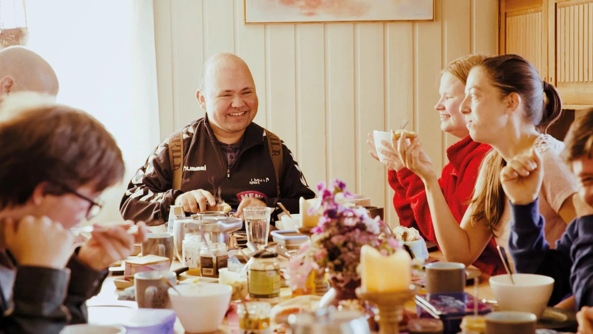 A smiling man sits a table full of food and crockery, flanked by other smiling people.