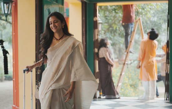 Promotional image for Too Desi Too Queer; two South Asian women in red dresses sit together in a field of long grass. One woman kisses the shoulder of the other woman while she rests on her knee.
