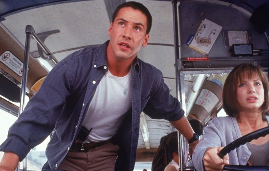 Keanu Reeves stands up in a bus and guides Sandra Bullock, who is sat at the wheel looking tense.