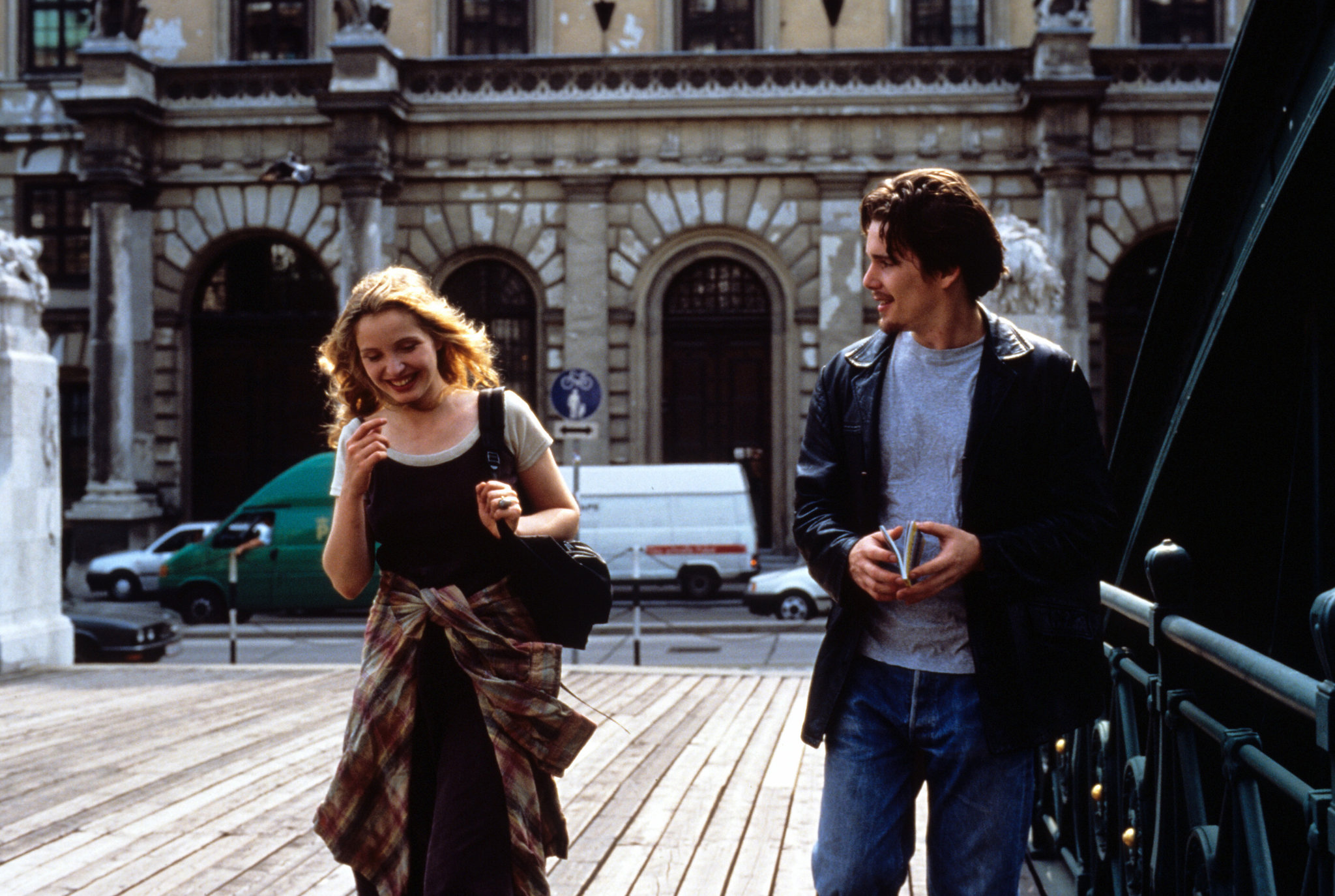 A still from Before Sunrise of Julie Delpy and Ethan Hawke walking side by side smiling at each other.