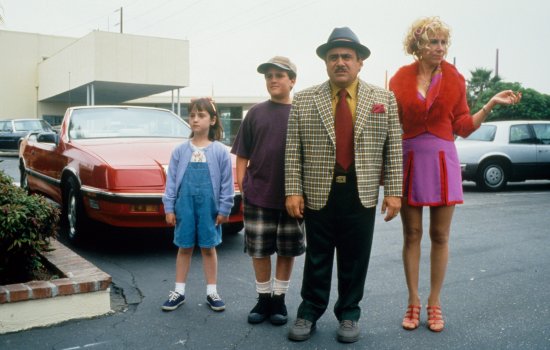 A still from Matilda [1996] featuring Matilda and the rest of her family stood in a parking lot wearing bright and colourful clothes.