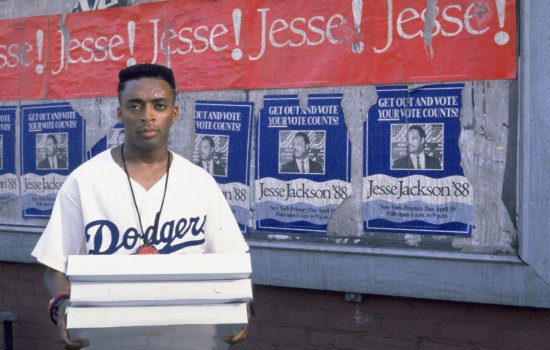 An image of Spike Lee stood holding pizza boxes in front of a wall plastered with Jesse Jackson '88 posters.