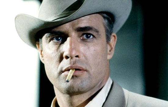 Marlon Brando stands smoking a cigarette, dressed in a suit and smart hat.