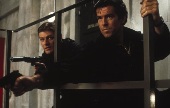 Pierce Brosnan and Sean Bean both point guns off screen, taking cover behind some bars.