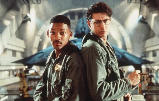 Will Smith and Jeff Goldblum stand side by side in an aircraft hangar, with a fighter jet in the background.