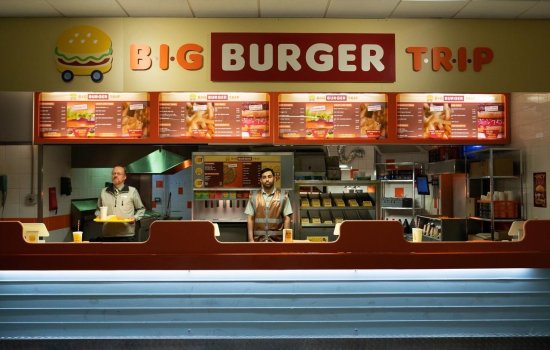 A young boy man stands alone at a burger restauarant late at night, staring out.