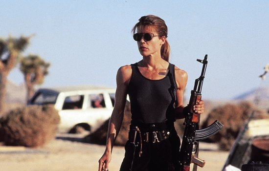 A still from Terminator 2 of Sarah Connor stood in a warm, desert location holding a machine gun.