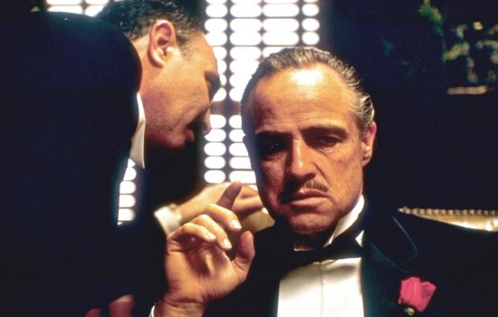 An iconic image from The Godfather of Marlon Brando as Don Corleone sat wearing a tuxedo as a man leans in and whispers in his ear.