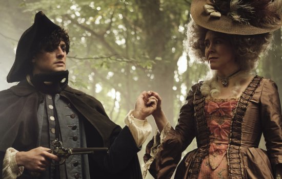 A man and woman in period costume hold hands in a wooded area.