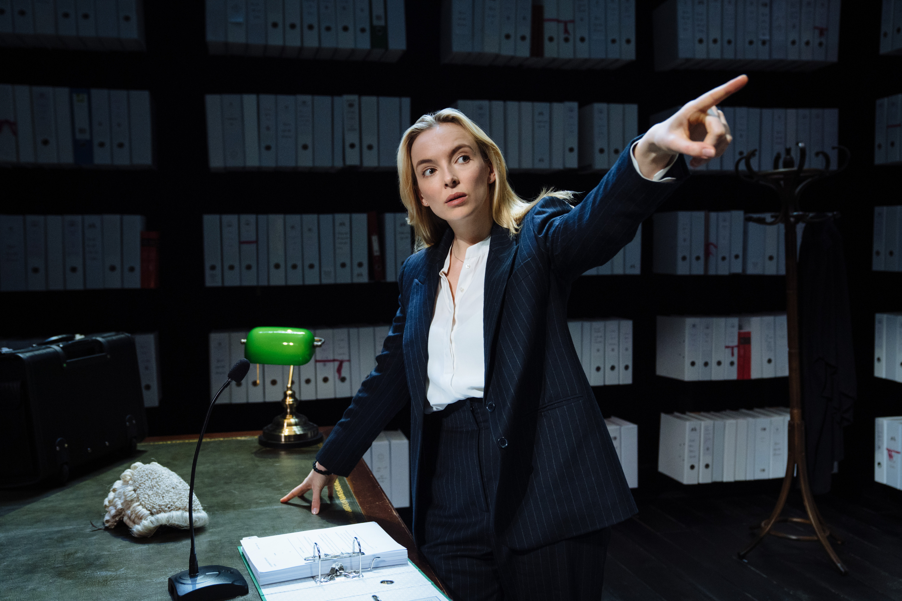 Jodie Comer stands at a desk, dressed in a suit and pointing off-screen.