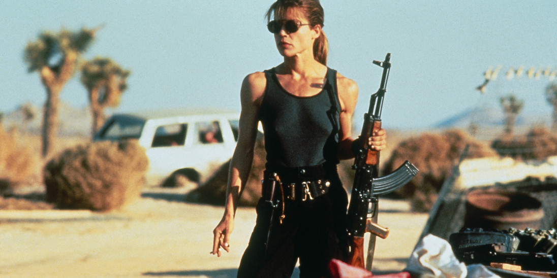 A still from Terminator 2 of Sarah Connor stood in a warm, desert location holding a machine gun.