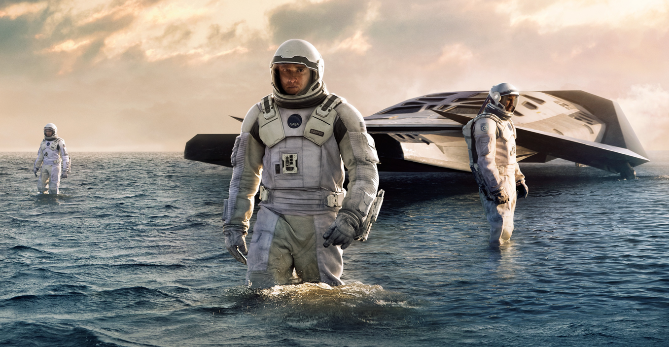 A group of people in spacesuits stand in the middle of water, their spaceship sits in the background.