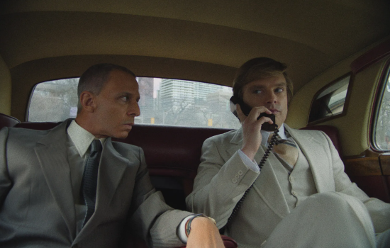 Two men in suits sit in a car, one if on a phone.