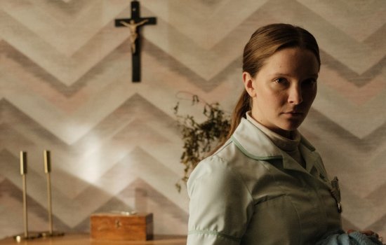 A woman stands in front of a crucifix displayed on a dark wall.