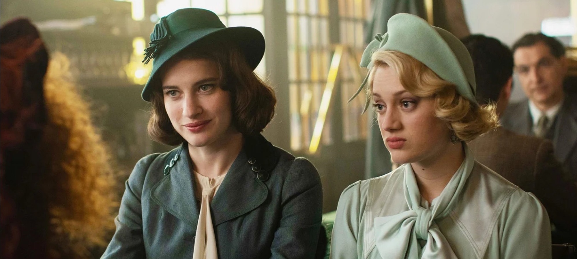 Two women sit side by side in 1930s costume. One smiles while one frowns.