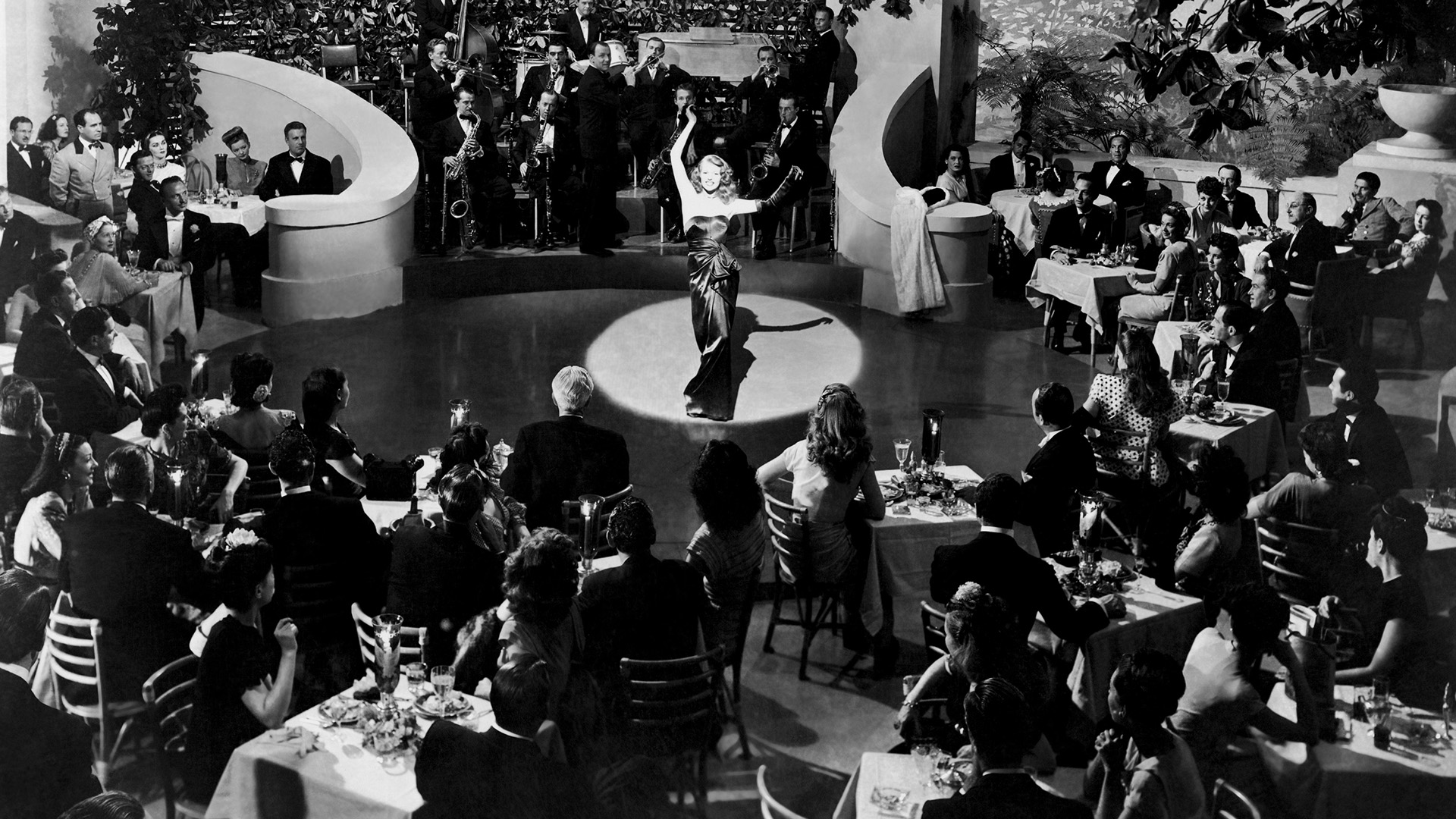 A black and white image of a woman stood lit by a spotlight in front of seated onlookers.