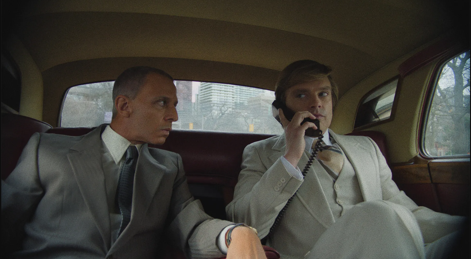 Two men in suits sit in a car, one if on a phone.