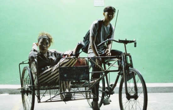 Two people side on a bike and cart next to a green background.