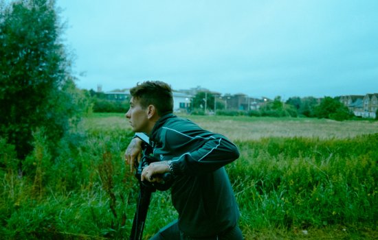 Barry Keoghan stands in a field leaning on a fence.