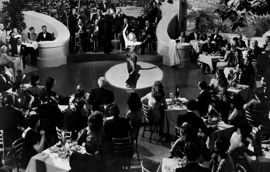 A black and white image of a woman stood lit by a spotlight in front of seated onlookers.